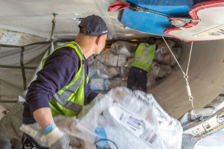 Alaska Airlines Ramp Service Agent (RSA) Carlos Arenas, foreground, passes a bag of mail to Lead RSA Metin Mehmedov. Both are working in the aft belly hold of the aircraft.