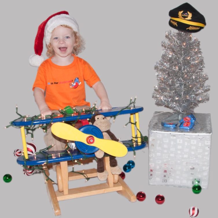 A young kiddo wearing a santa hat and rides an airplane rocking horse and poses next to a christmas tree topped with a captain's hat. 