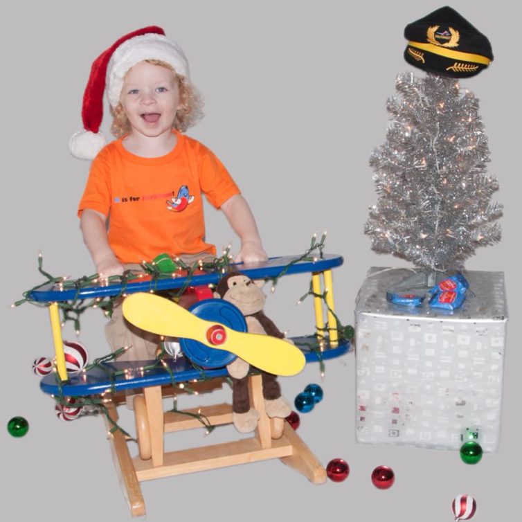 A young kiddo wearing a santa hat and rides an airplane rocking horse and poses next to a christmas tree topped with a captain's hat.