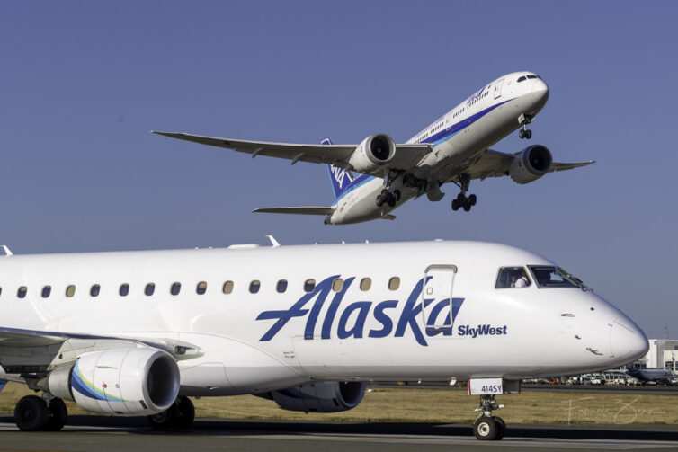 An Alaska Air Embraer waits on a taxiway at SEA while an ANA 787 takes off behind it