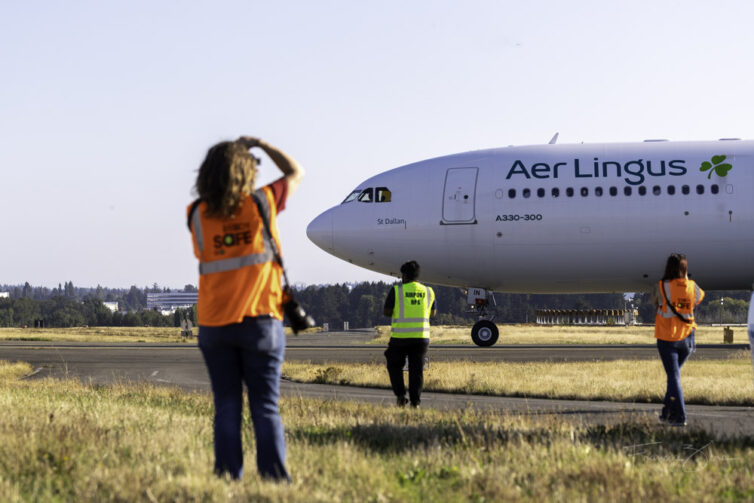 Several people standing in a grassy area taking photos of a passing airplane.